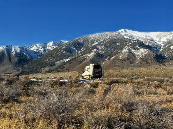 A light-colored rv rental van rests in a snow-dusted, brushy landscape with majestic, snow-capped mountains under a clear sky.. Custom Mercedes Benz Sprinter 2022