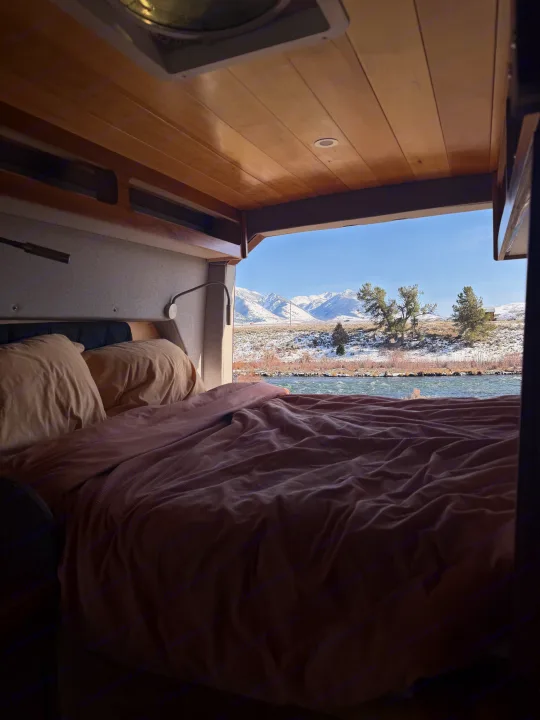 Scenic RV rental bedroom with cozy bed and wooden interior, offering panoramic views of snowy mountains and a flowing river.. Custom Mercedes Benz Sprinter 2022
