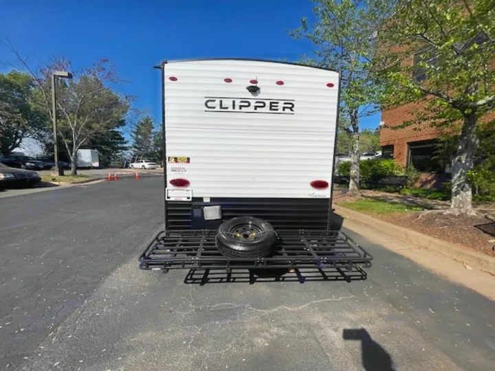 Rear view of a white Clipper travel trailer RV rental, equipped with a cargo carrier and spare tire in a sunny parking lot.. Coachmen Clipper 2025