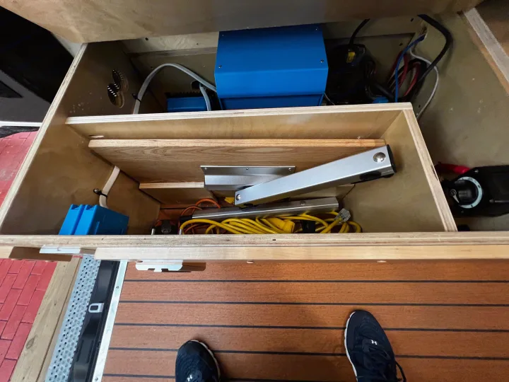 RV rental utility bay: blue electrical devices, wires, and storage organized within a wooden cabinet. Person's shoe visible on the floor.. Mercedes-Benz Sprinter 2023
