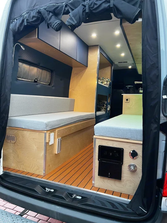 This inviting RV rental's camper van interior features light wood, grey cushions, bunk beds, and a slatted floor, seen from the rear.. Mercedes-Benz Sprinter 2023