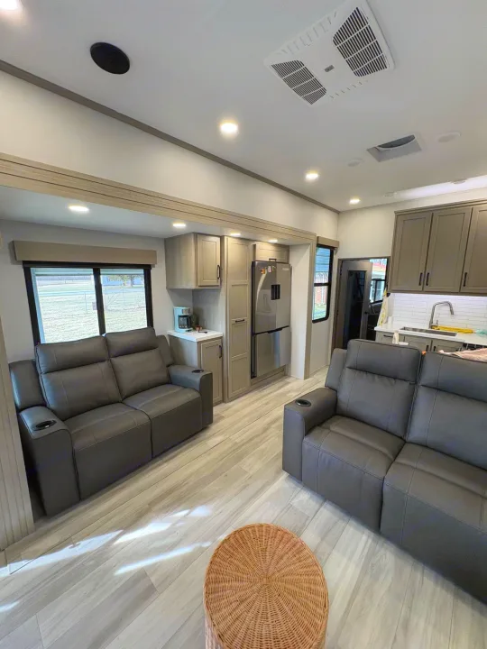 Spacious, modern RV rental interior: living area with comfy gray recliners, full kitchen, light wood floors, and white walls.. Grand Design Momentum M-Class Toy Hauler 2025