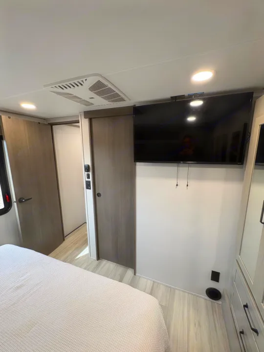 Inside a modern RV rental bedroom featuring a flat-screen TV, ceiling AC, light flooring, and comfortable bed.. Grand Design Momentum M-Class Toy Hauler 2025