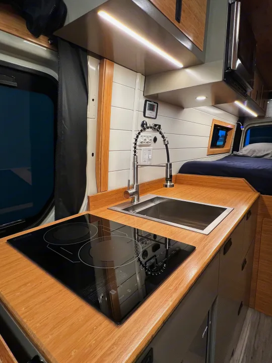 Modern RV rental kitchen with induction cooktop, sink, and cozy bed area, featuring bright overhead lighting.. Mercedes-Benz Sprinter 2024