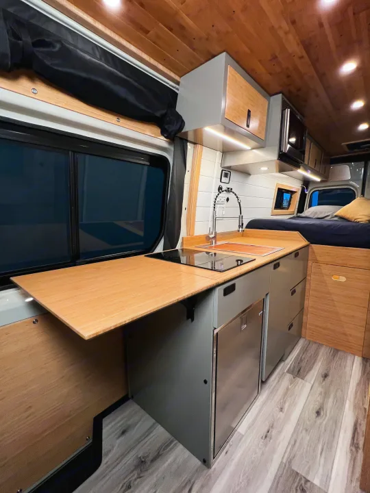 Enjoy this rv rental's modern interior. Wood kitchen with stove, sink, fridge & pull-out table. Cozy bed & sleek design.. Mercedes-Benz Sprinter 2024