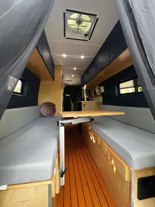 Modern RV rental interior: convertible dinette with gray cushions, wooden floor, overhead storage, and ceiling fan.. Mercedes-Benz Sprinter 2023