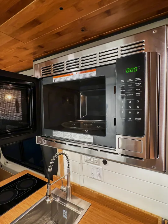 RV rental kitchen features an open microwave, sleek faucet, induction cooktop, and wooden ceiling.. Mercedes-Benz Sprinter 2024