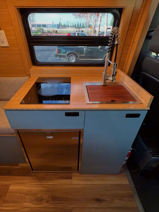 Sleek RV rental kitchen: bamboo counter, induction cooktop, sink with wooden cover, fridge, and window view outside.. Mercedes-Benz Sprinter 2024