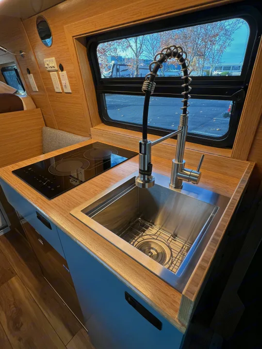 Modern RV rental kitchen featuring a stainless steel sink, a sleek pull-down faucet, and an induction cooktop on wooden countertops.. Mercedes-Benz Sprinter 2024
