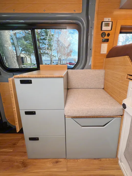 Modern **rv rental** interior: light gray storage, bamboo countertop, cushioned bench, and window with outdoor scenery.. Mercedes-Benz Sprinter 2024