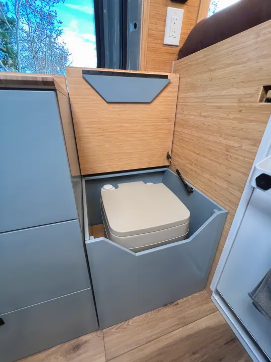 RV rental: A portable toilet revealed in a camper van's custom gray enclosure with an open bamboo lid.. Mercedes-Benz Sprinter 2024