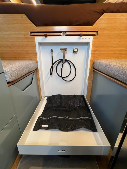 Compact RV rental shower/utility basin with pull-out hose, nozzle, and mesh bag, flanked by cushions and wood paneling.. Mercedes-Benz Sprinter 2024