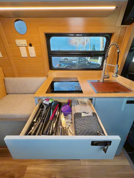 RV rental kitchen with open drawer showing organized utensils, induction cooktop, and sink. Cozy camper interior.. Mercedes-Benz Sprinter 2024
