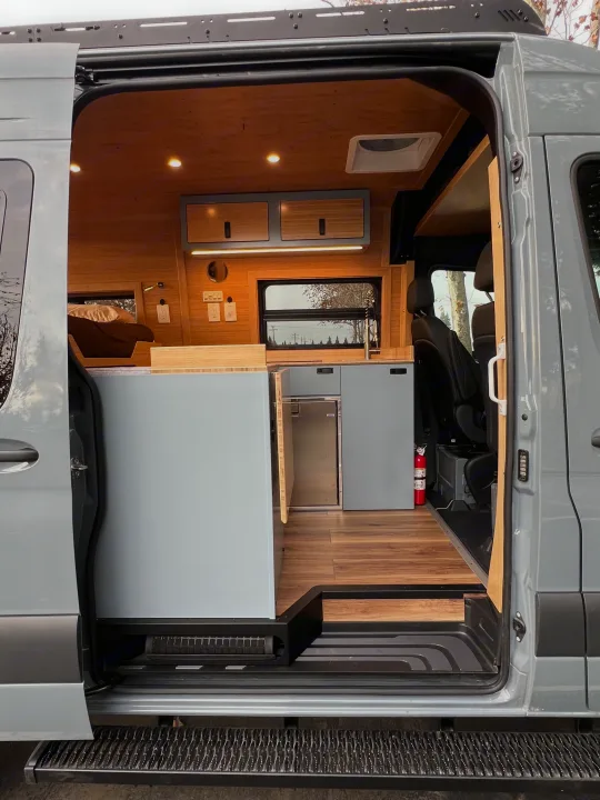 Step inside this custom rv rental camper van! Features a sleek kitchenette, cozy bed, and warm wooden interior, seen from the open side door.. Mercedes-Benz Sprinter 2024