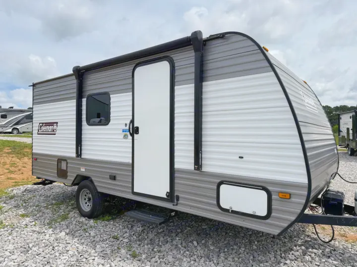 This grey and white Coleman travel trailer is parked on a gravel lot, ready for rv rental. It has a side door and awning.. Dutchmen Coleman 2024
