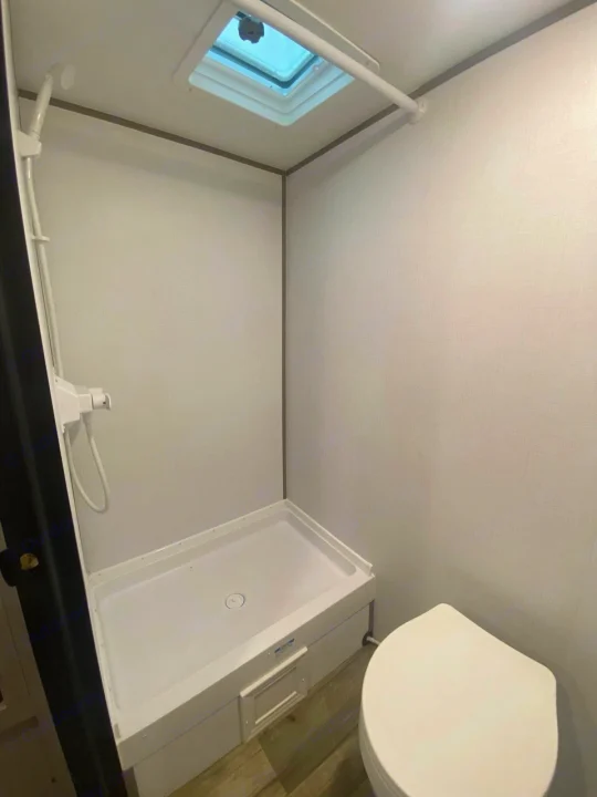 Compact RV rental wet bath with a white shower pan, handheld shower, toilet, and ceiling skylight. Practical for your travels.. Dutchmen Coleman 2024