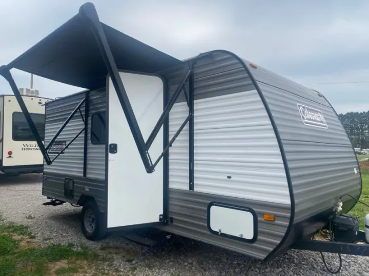 This grey and white Coleman travel trailer with its awning extended and door open is an ideal RV rental for your next trip.. Dutchmen Coleman 2024
