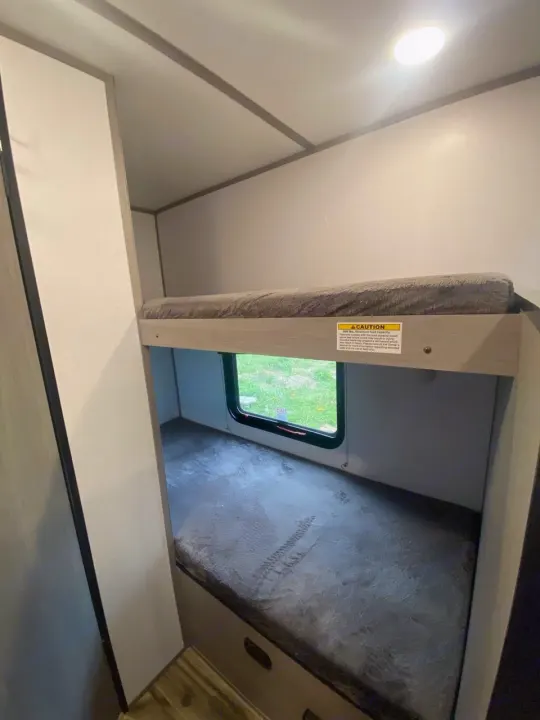 This RV rental features stacked bunk beds with mattresses and a window showing outdoor scenery. A cozy, compact sleeping area.. Dutchmen Coleman 2024