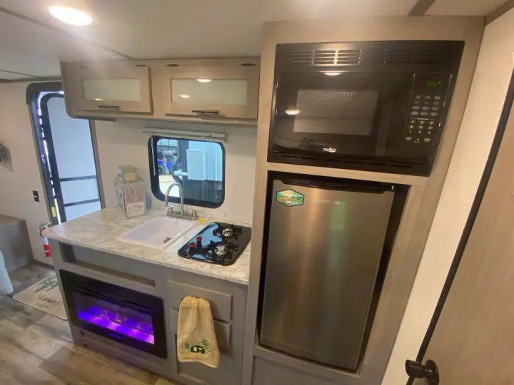 Compact RV rental kitchen features a microwave, mini-fridge, sink, 2-burner stove, and electric fireplace.. Dutchmen Coleman 2024