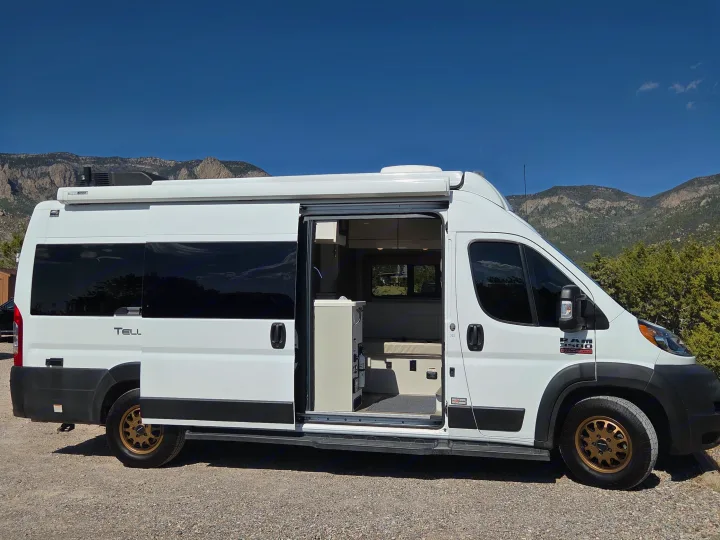 White Ram ProMaster rv rental campervan with its side door open, revealing the interior, against a mountain backdrop.. Thor Tellaro 2022