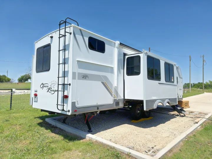 White Open Range rv rental with an extended slide-out and rear ladder, ready at a gravel campsite under clear blue skies.. Highland Ridge RV Open Range Roamer 2026