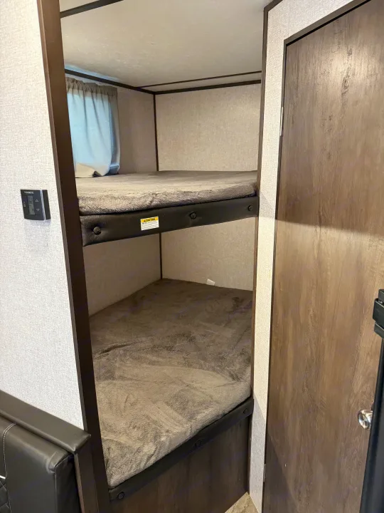 Cozy RV rental bunk beds with gray blankets, wall controls, and a wooden door, offering comfortable sleeping space.. Aspen Trail Aspen Trail Trailer 2021