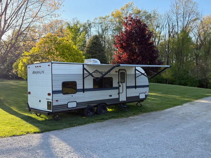 Ameri-Lite travel trailer (RV rental) set up on green grass with awning extended. Gravel drive and vibrant trees complete the outdoor camping scene.. Gulf Stream Ameri-Lite 2021