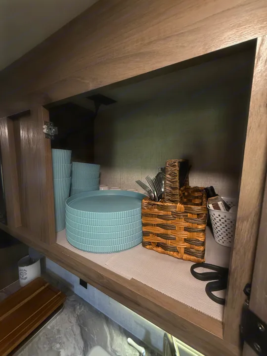 Organized RV rental kitchen cabinet with light blue ribbed dishes, stacked plates, cutlery, and storage baskets.. Gulf Stream Ameri-Lite 2021
