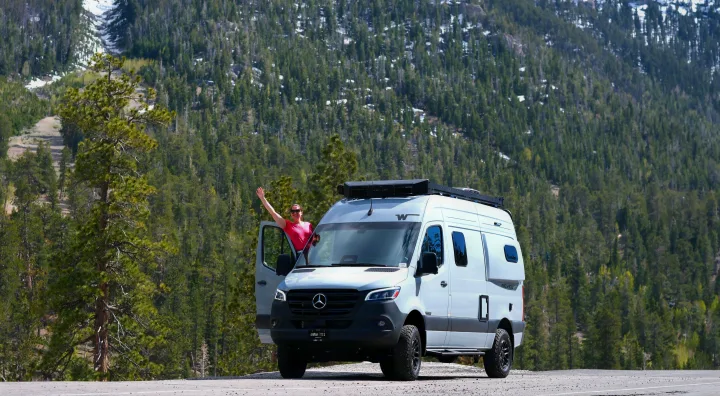 Woman in pink shirt waving from an rv rental Mercedes-Benz camper van on a mountain road amidst a dense forest.. Mercedes Winnebago 2026
