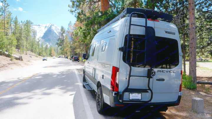 Grey Winnebago Revel rv rental camper van parked on a scenic mountain road. Snow-capped peak and pine trees fill the background.. Mercedes Winnebago 2026