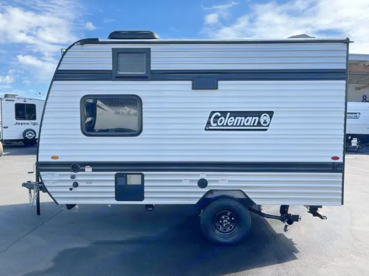 A small white Coleman travel trailer for rv rental, featuring black trim, windows, and a side logo, parked outdoors.. Keystone RV Keystone Coleman 13R 2026