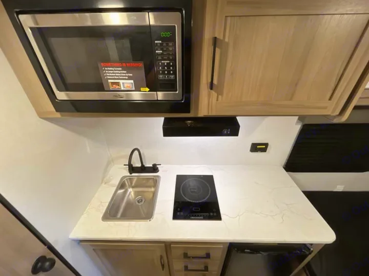 Compact RV rental kitchen with stainless microwave, sink, induction cooktop, and entertainment unit above a patterned countertop.. Keystone RV Keystone Coleman 13R 2026