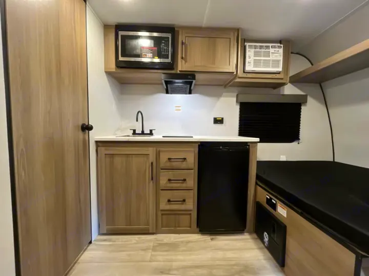 Modern rv rental interior: A compact kitchenette with microwave, sink, fridge, drawers, and an adjacent cozy bed.. Keystone RV Keystone Coleman 13R 2026