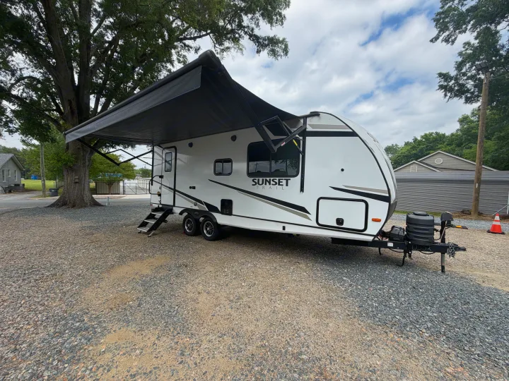A white Sunset Trail travel trailer RV rental with an extended awning is parked on a gravel lot under a large tree.. Crossroads RV Sunset Trail Super Lite 2024