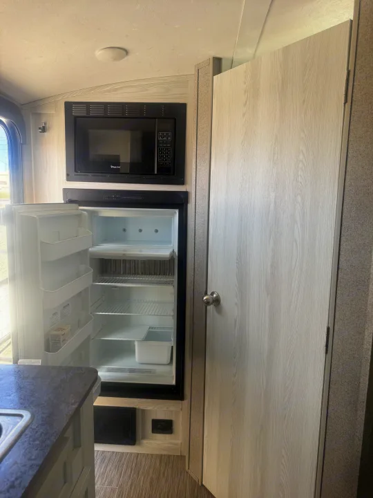 RV rental kitchen with a built-in microwave over an open refrigerator, next to a light wood door and counter.. Forest River Flagstaff E-Pro 2018