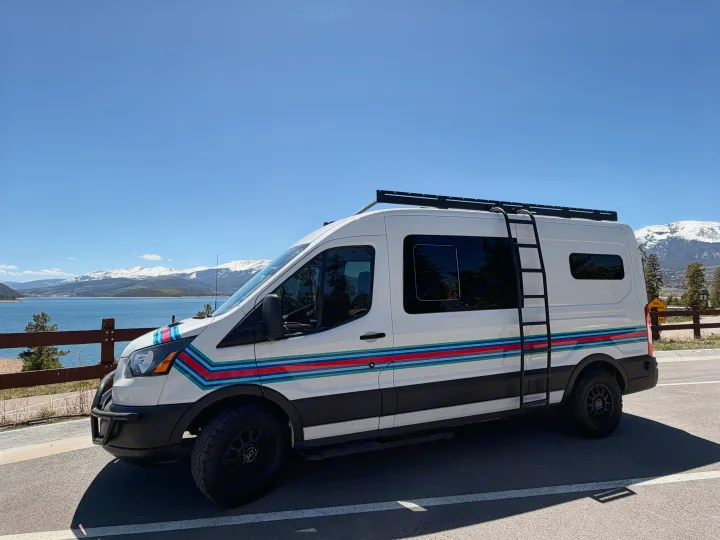 White RV rental with colorful stripes, roof rack, and ladder, parked by a scenic mountain lake with snowy peaks.. Ford Transit 2019