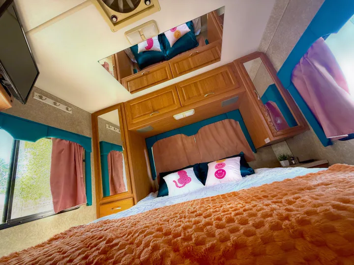 Vibrant rv rental bedroom with a fluffy orange bed, fun patterned pillows, teal curtains, and wooden cabinetry.. Four Winds 5000 2005