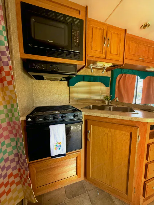 RV rental kitchen showing microwave, stove, oven, double sink, and light wood cabinets with a colorful curtain.. Four Winds 5000 2005