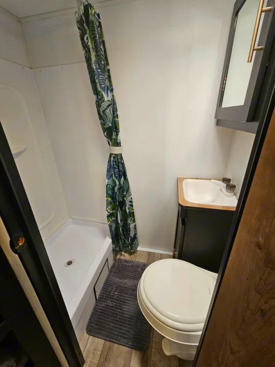 RV rental bathroom: Compact shower, toilet, sink, and mirror cabinet with a vibrant tropical curtain and dark bath mat.. Keystone RV Springdale 2022