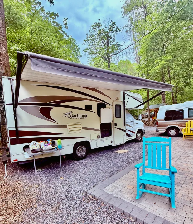 Enjoying a serene RV rental experience: a white Coachmen Freelander parked under lush trees with an extended awning, table, and blue rocking chair.. Coachmen Freelander 2015