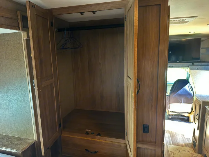 RV rental wardrobe: open wood closet with empty hangers & drawer. View into RV cockpit with a covered seat and TV.. Coachmen Freelander 2015