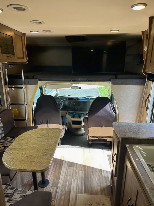View inside an RV rental showing the cab, overhead bunk bed with TV, a dinette, and a partial kitchen counter.. Coachmen Freelander 2015
