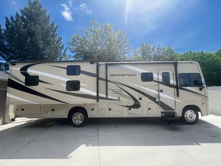 Exterior view of a beige and black Georgetown Class A RV rental, parked on a concrete driveway under a clear, sunny sky with trees.. Georgetown GT3 2020