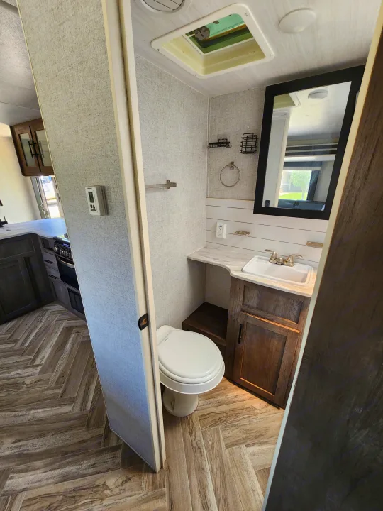 This RV rental features a modern bathroom with toilet, vanity, sink, and mirror, leading to a kitchen with herringbone flooring.. Forest River Wildwood X-Lite 2021