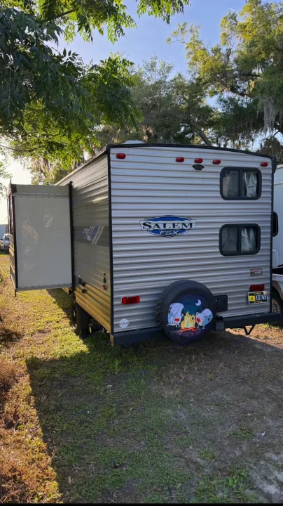 Salem FSX rv rental: silver travel trailer with slide-out extended, parked on grass under trees. Fun marshmallow spare tire cover.. Forest River Salem FSX 2022