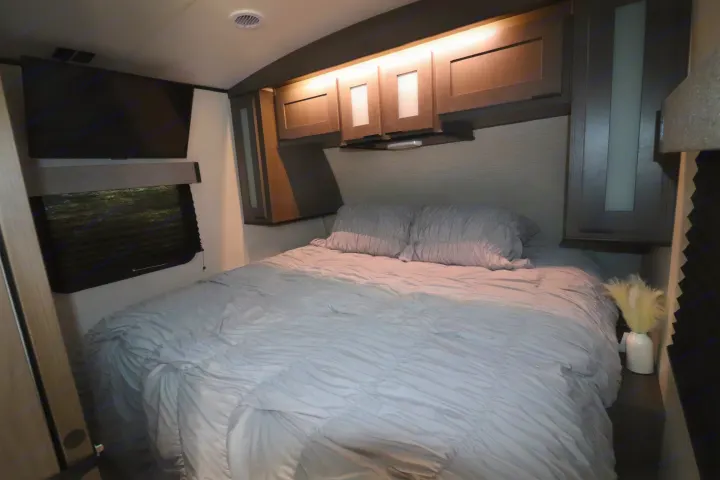 This RV rental bedroom offers a cozy bed with grey bedding, overhead lit cabinets, a wall TV, and a window with blind.. Cruiser RV MPG Ultra Lite 2021