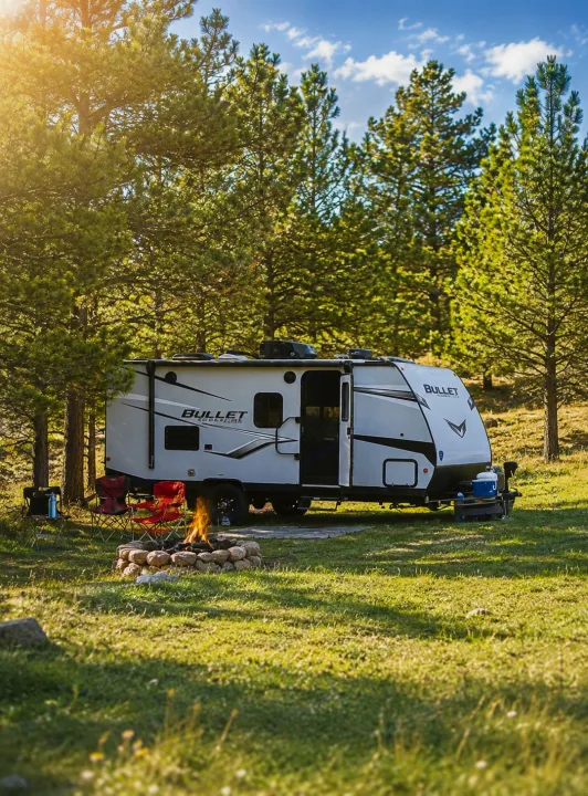 White RV rental parked in a sunlit forest campsite with a crackling campfire, chairs, and tall pine trees under a blue sky.. Keystone RV Bullet Crossfire 2022