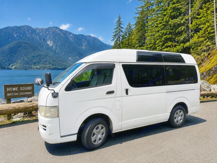 White RV rental van parked at Howe Sound, British Columbia, overlooking the water and forested mountains under a clear sky.. Toyota Hiace 2005