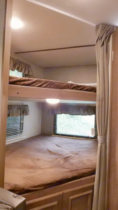 This RV rental bunk bed area features cozy brown blankets, windows with blinds, and a curtain, ideal for a comfortable stay.. Keystone RV Passport Ultra Lite 2019