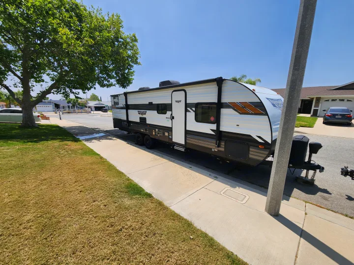 A white and black RV rental parked on a sunny residential street, with a large tree on the left and houses in the background.. Forest River Wildwood X-Lite 2021